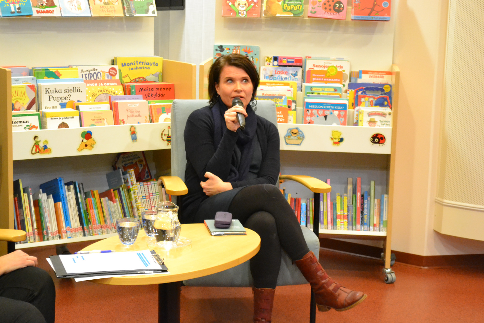 Marketta Pyysalo istuu mikrofoni kädessä. Taustalla kirjaston lastenkirjoja.